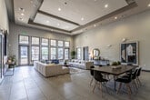 This thoughtfully designed single-floor community clubhouse offers a welcoming and multifunctional gathering space defined by soaring ceilings, clean architectural lines, and an open, airy layout. The main lounge area is a standout feature, showcasing dramatic ceiling height accentuated by decorative coffered beams and recessed lighting. Oversized windows and glass-paneled doors line the walls, flooding the interior with natural light and offering views of the surrounding greenery. The lounge is furnished with generously sized, channeled sectional sofas in neutral tones, complemented by bold navy accent chairs, layered area rugs, and a sculptural wooden coffee table — creating a refined yet comfortable social environment. A console table adorned with a round mirror and decorative accessories adds an elegant touch along one wall, alongside curated artwork. Transitioning into the adjacent area, wide-plank hardwood flooring sets the tone for a more active zone featuring round dining tables with vibrant yellow chairs and a stylish Sputnik-style chandelier overhead. This area flows seamlessly into a games and snack station, complete with a popcorn machine, a prep counter with sink, and casual seating around a dark-topped table. Sliding barn doors and arched passageways add architectural character throughout, tying the entire space together with a modern-meets-classic sensibility.