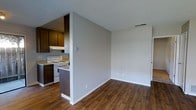 This single-floor residence offers a practical and functional layout designed for comfortable everyday living. The kitchen is equipped with a white electric range, a double-basin setup, and ample cabinetry finished in warm wood tones, providing generous storage and prep space. A peninsula-style counter serves as a natural divider between the kitchen and the adjacent living area, creating an open-concept flow that makes the space feel connected and airy. The living area is generously proportioned, with clean, neutral gray walls that provide a versatile backdrop for a variety of interior styles. Rich, dark hardwood-style flooring runs throughout, adding warmth and visual continuity across both areas. The textured ceiling adds a classic touch, while HVAC vents are strategically placed to ensure consistent climate control. White baseboards provide a crisp contrast against the flooring, contributing to a tidy, well-maintained aesthetic. A doorway visible on the far wall suggests additional rooms or access points beyond the main living space. Overall, the residence presents a clean, move-in-ready environment with a straightforward and efficient design.