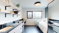 This thoughtfully designed single-floor studio unit offers a clean, modern aesthetic defined by a crisp white-and-black color palette throughout. The main living area seamlessly integrates sleeping, working, and kitchen functions into one efficient layout. A large window frames striking city skyline views, flooding the space with natural light and serving as a standout architectural feature of the room.

The kitchenette runs along one wall, featuring a dark countertop, white cabinetry with matte black hardware, a compact refrigerator, and a microwave on open wall-mounted shelving above. The counter extends to double as a functional desk area, complete with a chair, making it ideal for studying or working from home. A wall-mounted mini-split unit provides climate control throughout the space.

The sleeping area accommodates a full bed with built-in under-bed storage, complemented by a full-length mirror with a sleek black frame. A dedicated entry area provides a coat hook and digital access entry door, while an open doorway leads to the private bathroom. The bathroom is finished with large-format tile flooring, a toilet, and a tub-shower combination with a curtain rod, all accented with matte black fixtures. The overall ambiance is clean, contemporary, and efficiently organized.