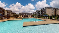 Located at 3050 Plantation Garden Dr in Fayetteville, North Carolina, this outdoor amenity area serves as a central gathering space for the community. The centerpiece is a generously sized swimming pool with clear, blue water, surrounded by a wide concrete deck lined with contemporary lounge chairs, offering ample space for sunbathing and relaxation.

Adjacent to the pool is a well-appointed single-story clubhouse pavilion characterized by a prominent gabled roofline, white columnar supports set atop stacked stone bases, and board-and-batten siding. The covered porch area extends the living space outdoors and features a floor-to-ceiling stacked stone fireplace with a rustic wood mantel, creating a warm and inviting focal point. Comfortable seating, including a plush sofa and lounge chairs, is arranged throughout the covered patio, encouraging social gatherings in any season.

The stamped concrete flooring of the pavilion adds a refined aesthetic detail, while the surrounding landscaping provides greenery and color that soften the hardscape. A wrought-iron perimeter fence encloses the pool area, enhancing both safety and visual definition. Views of the surrounding multi-story residential buildings are visible from the pool deck, reinforcing the community-centered character of this amenity space.