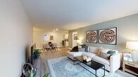 This thoughtfully designed 1-bedroom, 2-bathroom residence offers a spacious and functional single-floor layout. The open-concept living and dining area is anchored by light wood-style flooring, recessed lighting, and a soothing neutral color palette accented by sage green walls. A large sectional sofa, patterned area rug, and natural light from generous windows create a warm, inviting atmosphere in the living room. The galley-style kitchen features white shaker cabinetry, light quartz countertops, and stainless steel appliances, opening seamlessly into the dining area where a round wood table and modern chairs offer an elegant setting. The primary bedroom is a comfortable retreat with plush carpeting, a bold teal accent wall, and natural light from a window. It is complemented by a well-organized walk-in closet with built-in shelving and hanging rods. Two private office spaces provide dedicated areas for work or study, each finished with carpet flooring and neutral walls. Both bathrooms are clean and bright, featuring wood-look tile flooring, white vanities, and understated finishes. Three total closets ensure exceptional storage throughout. The overall ambiance blends contemporary style with practical livability.
