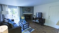 This well-appointed single-floor space combines comfort and style in a thoughtfully designed layout. The living area serves as the centerpiece, showcasing rich, dark hardwood flooring that anchors the room with warmth and character. A corner fireplace with a clean white mantel provides a cozy focal point, complemented by a striking metal bird wall sculpture that adds an artistic touch to the accent wall. Natural light pours in generously through a large sliding glass door dressed with white curtains, offering a view of a private outdoor area with greenery beyond. The space is furnished with a plush navy sofa and a coordinating blue accent chair, arranged around a glass-topped coffee table that rests on a geometric area rug, creating an inviting conversational arrangement. A dark wood media console positioned opposite the seating area provides both storage and display space. The neutral gray and white wall palette allows the carefully chosen décor and furnishings to take center stage. Overall, the space exudes a relaxed yet polished ambiance, blending functional design with tasteful decorative details that make it feel both livable and refined.