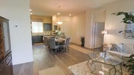 This single-story home offers an inviting open-concept design that connects the main living spaces in a cohesive and functional layout. The kitchen is equipped with rich dark cabinetry, light stone countertops, and a full suite of appliances, complemented by a window that brings in natural light. A decorative valance adds a warm, curated touch above the window. The dining area sits at the heart of the open floor plan, centered around a round wooden dining table with upholstered chairs and illuminated by a classic pendant light overhead. Recessed lighting throughout the space ensures consistent, comfortable illumination. Wide-plank hardwood-style flooring flows seamlessly across the main living areas, adding warmth and visual continuity. The neutral, earthy wall tones create a calm and welcoming ambiance that pairs well with the darker furnishings. A glimpse into the adjacent rooms reveals additional living and utility spaces, suggesting a well-rounded and practical floor plan. Tasteful decorative accents, including fresh florals and potted greenery, contribute to the overall polished and livable character of this home.