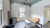 This single-floor space showcases an industrial loft aesthetic defined by whitewashed exposed brick walls and elevated ceilings accented with exposed structural beams painted in a cool grey tone. Track lighting runs along the ceiling, providing adjustable, directed illumination throughout the room. The painted concrete flooring adds to the raw, urban character of the space.

A standout feature is the large window fitted with a cellular shade blind, which frames a striking view of a city bridge and waterway beyond — offering an urban backdrop that adds visual depth and natural light to the interior. The window's placement draws the eye and serves as a natural focal point within the room.

The living area is furnished with a tan leather futon sofa and an additional seating piece, centered around a minimalist white rectangular coffee table. A dark multi-drawer chest sits against one wall, providing practical storage. The overall atmosphere is open and versatile, blending industrial architectural character with functional, casual furnishings — making it a flexible space well-suited for both relaxing and entertaining.