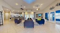 Located at 917 Country Club Park in DeLand, Florida, this single-story community clubhouse offers a well-appointed gathering space designed for both comfort and functionality. The main lounge area is the heart of the facility, featuring soaring vaulted ceilings with a dramatic skylight that floods the room with natural light. Dual ceiling fans, recessed lighting, and large French doors leading to the outdoors enhance the bright, airy ambiance. The lounge is furnished with plush blue sofas and accent chairs arranged around a central rug, creating an inviting setting for socializing.

Adjacent to the lounge is a full kitchen complete with white cabinetry, granite countertops, a breakfast bar with ornate bar stools, and stainless steel appliances — ideal for community events and gatherings. A dedicated office area with glass partitions provides a functional workspace visible from the main lounge.

A separate fitness room, finished in bold blue, houses an array of exercise equipment and is accessible directly from the main lounge through French doors. The entryway features elegant French doors with a striking arched transom window, flanked by patterned accent chairs, offering a welcoming first impression. Throughout, large-format tile flooring ties the space together, creating a clean, cohesive aesthetic suited for an active community lifestyle.