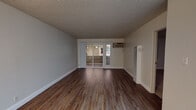 Located at 3777 Mowry Ave in Fremont, California, this single-floor unit offers a practical and well-maintained layout across several distinct living spaces. The open living area is finished with rich, dark hardwood-style flooring and features a textured ceiling, neutral gray walls, and a wall-mounted HVAC unit. Sliding glass doors at the far end of the living space provide access to an outdoor area, allowing natural light to filter in and offering a connection to the exterior surroundings. The kitchen is efficiently designed with white cabinetry, light granite-style countertops, and white appliances including an electric range and refrigerator. Light wood-style flooring and under-cabinet lighting give the kitchen a clean, functional aesthetic. A bold red entry door adds a distinctive accent. The bathroom is neatly appointed with a white vanity, a full bathtub and shower combination, wood-style flooring, and crisp white finishes throughout. The bedroom is carpeted and features a sliding window that looks out onto the surrounding neighborhood, providing ventilation and natural light. Additional interior spaces include a hallway with a closet and a wall heater unit. Overall, the unit presents a clean, move-in ready environment with consistent finishes throughout.
