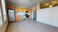 Located at 5800 Highlands Plaza Dr, this single-floor unit offers a clean, modern aesthetic with an open-concept layout that seamlessly connects the kitchen and living areas. The kitchen is well-appointed with warm maple-toned cabinetry providing generous storage, complemented by a full suite of stainless steel appliances including a refrigerator, microwave, electric range, and a stainless sink. Light-toned luxury vinyl plank flooring extends throughout the space, adding durability and a cohesive, contemporary feel underfoot. Track lighting on the kitchen ceiling ensures functional illumination for food preparation, while the open layout allows natural light to flow through the living area. The neutral white walls create a bright, airy atmosphere that lends itself to a variety of interior styles. A corridor visible from the main living space leads to additional rooms, suggesting a practical and well-organized floor plan. The overall ambiance is modern and move-in ready, with thoughtful design details that balance both style and functionality throughout the unit.