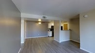 This single-floor space showcases an open-concept design that seamlessly connects the living area and kitchen, creating a functional and cohesive layout. The living area features smooth hardwood-style flooring in a warm neutral tone, complemented by light gray walls and white baseboards that lend a clean, contemporary feel. A ceiling fan with a built-in light fixture is centrally positioned, providing both airflow and ambient lighting. Overhead, a ceiling-mounted HVAC vent ensures efficient climate control throughout the space.

The kitchen is situated toward the rear and is equipped with white cabinetry, black appliances including a range and microwave, and a dark tile backsplash that adds visual contrast. A pass-through bar counter connects the kitchen to an adjacent dining or breakfast nook area, which features a warm yellow accent wall, offering a bright and distinct space for casual dining.

A doorway visible on the left side of the living area suggests access to additional rooms. Overall, the space presents a practical and well-maintained interior with a neutral palette, making it a versatile and move-in ready environment.