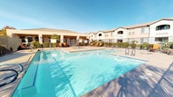 This outdoor amenity area is designed to support relaxed, resort-style living within a multi-building residential community. The centerpiece is a generously sized swimming pool with clear blue water, complemented by an adjacent spa that features bubbling jets—both enclosed by a secure iron fence and surrounded by a wide concrete deck outfitted with lounge chairs for sunbathing. The pool area benefits from abundant natural sunlight and open sky views, creating a bright and inviting atmosphere throughout the day.

A covered patio lounge area provides a shaded retreat alongside the main building. Supported by substantial stucco columns, the structure shelters a seating arrangement of wicker furniture with bold orange cushions, offering a comfortable space to gather or unwind. French doors connect this lounge directly to the interior, enhancing accessibility and flow between indoor and outdoor spaces.

A dedicated BBQ station, equipped with a large stainless steel multi-burner grill, is positioned among decorative gravel landscaping and manicured shrubs, making it well-suited for outdoor cooking and social gatherings. Throughout the space, lush greenery, palm trees, and low-maintenance desert plantings frame the amenities, contributing to a well-maintained, cohesive aesthetic.