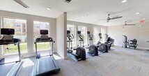 Located in Grand Prairie, Texas, this well-appointed fitness center is designed to support a full range of workout routines in a bright, modern environment. The single-floor layout is organized into distinct functional zones, each thoughtfully equipped and finished with neutral-toned carpet tile flooring and crisp white walls throughout.

The cardio zone is lined with large windows that flood the space with natural light and offer views of the surrounding community. This area houses a row of treadmills, elliptical trainers, and cycling machines, all positioned to take advantage of the outdoor views. Ceiling fans overhead provide additional air circulation, enhancing comfort during use.

A dedicated strength training area features a multi-station cable and weight machine, a flat bench, and a selection of free weights, complemented by wall-mounted mirrors that visually expand the space and allow users to monitor their form. A wall-mounted flat-screen television adds an entertainment element to the workout experience.

The facility also includes a separate stretching and functional fitness room, equipped with stability balls and floor-to-ceiling mirror panels. This versatile space is ideal for yoga, Pilates, or warm-up routines, and provides direct access to the outdoor area via glass-panel doors. The overall ambiance is clean, open, and motivating — designed to accommodate a variety of fitness needs.