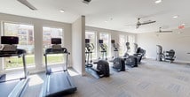 Located in Grand Prairie, Texas, this well-appointed fitness center is designed to support a full range of workout routines in a bright, modern environment. The single-floor layout is organized into distinct functional zones, each thoughtfully equipped and finished with neutral-toned carpet tile flooring and crisp white walls throughout.

The cardio zone is lined with large windows that flood the space with natural light and offer views of the surrounding community. This area houses a row of treadmills, elliptical trainers, and cycling machines, all positioned to take advantage of the outdoor views. Ceiling fans overhead provide additional air circulation, enhancing comfort during use.

A dedicated strength training area features a multi-station cable and weight machine, a flat bench, and a selection of free weights, complemented by wall-mounted mirrors that visually expand the space and allow users to monitor their form. A wall-mounted flat-screen television adds an entertainment element to the workout experience.

The facility also includes a separate stretching and functional fitness room, equipped with stability balls and floor-to-ceiling mirror panels. This versatile space is ideal for yoga, Pilates, or warm-up routines, and provides direct access to the outdoor area via glass-panel doors. The overall ambiance is clean, open, and motivating — designed to accommodate a variety of fitness needs.