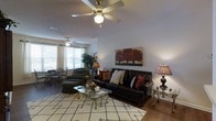This single-story space centers around a thoughtfully arranged living area that combines comfort and style. The room features hardwood flooring throughout, complemented by a large, patterned area rug that anchors the seating arrangement. A dark leather sofa with nailhead trim serves as the focal point, adorned with an assortment of decorative pillows in warm, earthy tones. A matching upholstered chair and ottoman provide additional seating, creating an inviting conversational layout.

Two ceiling fans are installed throughout the space, contributing to both airflow and ambiance. Natural light filters in through multiple windows dressed with curtains, brightening the neutral-toned walls. A vibrant autumn landscape painting adds a bold decorative element above the sofa, echoing the warm color palette of the room.

A glass-top coffee table with ornate gold detailing sits at the center of the seating area, while a side table with a classic table lamp adds layered lighting. In the background, a dining area with wrought-iron chairs is visible near the windows, alongside a lush indoor plant that adds a natural, organic touch to the overall setting.