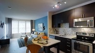This thoughtfully designed single-floor space showcases a seamless open-concept layout that integrates the kitchen, dining, and living areas into one cohesive and functional environment. The kitchen is outfitted with sleek dark cabinetry, light quartz countertops, and a striking mosaic tile backsplash that adds texture and visual interest. A stainless steel sink and modern fixtures complement the well-appointed cooking space, which also features stainless steel appliances. A compact dining area with a dark table and contemporary wooden chairs sits adjacent to the kitchen, providing an intimate space for meals. The living area is anchored by a bold teal accent wall, which adds a pop of color and depth to the room. A comfortable sofa with decorative pillows, a floor lamp, and thoughtfully placed artwork contribute to a warm and inviting atmosphere. Track lighting along the ceiling provides adjustable illumination throughout the open space, while a large window in the living area allows natural light to fill the room. The flooring transitions from light hardwood in the kitchen and dining zones to a patterned area rug in the living space, clearly defining each functional area while maintaining an open, airy feel throughout.