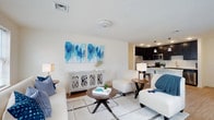This well-appointed single-floor residence offers a thoughtfully designed open-concept layout that connects the living and kitchen areas in a seamless, functional flow. The living area is bright and welcoming, featuring light-toned walls, smooth white ceilings with recessed ventilation, and warm hardwood-style flooring that adds depth and warmth to the space. A large, decorative triptych of blue abstract artwork serves as a striking focal point above a distressed white console table, lending the room a coastal-inspired charm. The space comfortably accommodates a plush seating arrangement centered around a round wooden coffee table and anchored by a soft area rug with a subtle geometric pattern. Just beyond the living area, the kitchen stands out with rich dark cabinetry, pendant lighting, and stainless steel appliances, including a microwave and range. A kitchen counter with bar seating creates a natural transition between the two areas, ideal for casual dining or entertaining. The overall ambiance is clean, contemporary, and inviting, with a cohesive color palette of whites, blues, and warm wood tones that creates a relaxed yet polished atmosphere throughout the home.