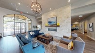 Located at 8823 Dugas Rd in San Antonio, TX, this single-story community clubhouse offers a thoughtfully designed layout with a variety of amenities. The centerpiece is a grand lounge area featuring rich hardwood flooring, soaring ceilings, and an eye-catching orb chandelier. A dramatic floor-to-ceiling natural stone fireplace wall anchors the room, while large arched French doors flood the space with natural light and provide views of the surrounding landscape. Multiple seating arrangements with blue velvet sofas and upholstered chairs create an inviting, relaxed atmosphere. Adjacent to the lounge, a catering kitchen is outfitted with granite countertops, stainless steel appliances, a subway tile backsplash, and a large window overlooking an outdoor covered area. A coffee and beverage bar, accented with deep blue mosaic tile, provides a cozy gathering spot alongside high-top dining tables. The business center offers a quiet workspace with a built-in desk station for multiple users, curated artwork, and comfortable lounge seating. A game room with foosball and framed artwork adds a recreational element. The expansive fitness center is equipped with a full range of cardio and strength machines, multiple flat-screen TVs, and large windows with garden views, completing this versatile and well-rounded community facility.