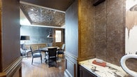 This distinguished single-floor space masterfully blends classic architectural detail with a moody, refined aesthetic. The centerpiece of the room is a stunning ornate tin ceiling, featuring intricate embossed medallion patterns that draw the eye upward and lend a sense of historic grandeur. Rich, warm-toned wood wainscoting and substantial column accents frame the walls, providing elegant contrast against the deep slate-blue upper wall surfaces that give the space a sophisticated, dramatic character.

The dining or conference area is anchored by a dark wood table surrounded by traditional upholstered chairs, all bathed in warm light from an industrial-style pendant fixture that complements the room's layered design sensibility. Wide-plank hardwood floors run throughout, adding warmth and continuity underfoot.

A built-in cabinetry section features a striking marble countertop with bold veining, offering both functional surface space and a luxurious material accent. A wood-framed doorway provides a natural transition point within the layout, maintaining the cohesive, classically inspired design language throughout. Overall, the space presents a compelling blend of ornate detailing and dark, intimate tones that create an atmosphere of elegance and refinement.