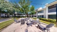 Located at Lakepointe Landing in Plantation, Florida, this outdoor communal lounge area offers a relaxed, resort-inspired atmosphere designed for social gathering and leisure. The centerpiece of the space is a fire pit encircled by a generous arrangement of gray Adirondack chairs, all set atop a sandy surface that lends a casual, beach-like feel to the environment. Overhead, strands of globe string lights are strung between tall poles, creating a warm and inviting ambiance during evening hours. Mature, leafy trees provide generous natural shade throughout the area, contributing to a cool and comfortable outdoor retreat even under Florida's sunny skies. The lush green lawn surrounding the sandy zone adds to the verdant setting, while well-maintained shrubbery borders the perimeter. The backdrop features a two-story residential building with balconies and large windows, integrating the communal space seamlessly within the broader community grounds. The open layout encourages residents to gather, unwind, and connect in a thoughtfully designed outdoor setting that balances natural beauty with functional amenities.