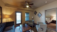 This single-floor space offers a comfortable and cohesive living environment defined by its warm, neutral color palette and thoughtful design details. The living area is anchored by rich dark hardwood flooring that runs throughout, providing a grounding contrast to the soft beige walls. A ceiling fan with a built-in light fixture serves as a functional focal point overhead, ensuring year-round comfort and air circulation. The seating arrangement features upholstered chairs in a muted teal-gray tone, accented with nailhead trim, complemented by a rustic wooden coffee table that adds warmth and texture to the space. Decorative framed artwork on the walls adds a personalized touch, while a geometric sculptural accent piece on the coffee table contributes visual interest. Natural light filters into the room through a window fitted with horizontal blinds, and a glass-panel door opens directly to what appears to be a balcony or exterior walkway, further brightening the interior. The overall ambiance is relaxed yet stylish, blending functional furnishings with tasteful décor to create an inviting and livable space suited for everyday use.