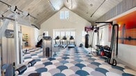 This fitness center is a standout space defined by its dramatic vaulted ceiling clad in whitewashed shiplap, which rises to an impressive peak and creates an open, airy atmosphere throughout. Track lighting runs along the ceiling lines, complementing the generous natural light that pours in through multiple large windows and glass French doors, offering views of the surrounding outdoor landscape, including palm trees and neighboring scenery.

The floor is one of the space's most distinctive design features — a bold, scallop-patterned carpet in shades of blue, gray, and white that adds a coastal, energetic character to the interior. The main workout area is generously sized and thoughtfully arranged to accommodate a wide variety of fitness activities. Cardio equipment, including treadmills and elliptical machines, lines the perimeter near the windows, allowing users to exercise while enjoying the views. A dedicated strength and functional training zone includes a cable machine, a punching bag, battle ropes, a pull-up rig, barbells, weight plates, and a full dumbbell rack. A mounted flat-screen television is positioned for easy viewing during workouts. An additional nook area provides space for stretching, balance equipment, and supplementary fitness accessories. The combination of architectural elegance and comprehensive equipment makes this a truly inspiring place to train.