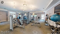 This fitness center at 108 Shadowood Drive in Chapel Hill, NC offers a comprehensive and well-appointed exercise facility designed to support a wide range of workout routines. The single-floor layout provides generous, open floor space that comfortably accommodates a diverse collection of equipment without feeling cramped.

The cardio zone features multiple treadmills, elliptical trainers, and stationary bikes from reputable brands, all positioned along windowed walls that allow natural light to filter in and provide views of the surrounding greenery. The strength training area is equally well-equipped, with cable and pulley machines, weight benches, a leg press machine, a rowing machine, and a selection of medicine balls and balance tools such as a BOSU ball.

The facility is finished with tasteful, neutral-toned carpeting and soft gray walls, lending a clean and inviting atmosphere. Tray ceilings with elegant pendant and flush-mount lighting elevate the aesthetic, giving the space a polished, upscale feel. A lobby-style anteroom provides a welcoming transition into the gym, complete with a tray ceiling, vending machines for convenient refreshments, and French doors leading to an adjacent lounge area with seating. A wall-mounted television adds entertainment options for those engaging in longer cardio sessions. The overall ambiance is bright, functional, and well-maintained.