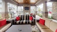 The Outdoor Lounge is a well-appointed covered gathering space designed to blend comfort and functionality in an open-air setting. The structure is defined by stacked-stone columns and accent walls that lend a refined, natural aesthetic throughout. A rich wood-planked ceiling with recessed lighting anchors the covered portion of the lounge, creating a warm and inviting atmosphere even after sunset.

At the heart of the space, a full stacked-stone fireplace serves as a dramatic focal point, topped by a large wall-mounted flat-screen television — ideal for entertaining or relaxed viewing. Multiple seating arrangements featuring wicker-frame sofas and chairs with bold striped and neutral cushions accented by red throw pillows are arranged around area rugs, encouraging conversation and relaxation.

An outdoor kitchen equipped with a gas grill and prep area provides ample space for cooking and hosting. Herringbone-patterned brick pavers extend throughout the lounge floor, adding texture and visual interest underfoot. Open on the sides, the space allows for natural breezes and views of the surrounding greenery and community, maintaining a seamless connection between the sheltered lounge and the outdoors.