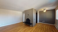This single-floor space is defined by a standout architectural centerpiece: a floor-to-ceiling painted brick fireplace with an arched iron door and an integrated bench, anchoring the open living area with character and warmth. Hardwood floors extend throughout the main living spaces, complementing the clean white walls and adding a warm, classic feel. Natural light filters in through windows fitted with plantation shutters near the entryway, brightening the open layout. A ceiling fan provides additional comfort in the adjacent dining or secondary living area, which features contrasting gray-painted walls.

The kitchen is well-equipped, showcasing rich dark wood cabinetry, stone-look granite countertops, a stainless steel sink with a modern pull-down faucet, a tiled backsplash with a decorative mosaic accent border, and black appliances. The space offers generous counter and storage space for everyday use.

The bathroom features large-format earth-toned tiles covering the walls, accented by a mosaic tile border, surrounding a full soaking tub with a frosted privacy window above. The bedroom is a calm, neutral retreat with plush carpeting, a baseboard heating unit, and a window overlooking the exterior. The overall layout is functional and cohesive, blending classic architectural details with practical, updated finishes throughout.