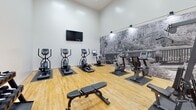 Located at 1301 Buckingham Station Dr in Midlothian, Virginia, this two-floor community facility is designed to support both active and social lifestyles. The fitness area is a standout, boasting soaring double-height ceilings and hardwood-style flooring throughout. It is comprehensively equipped with a multi-station strength training machine, a rack of dumbbells in varied weights, treadmills, ellipticals, upright and recumbent stationary bikes, and a selection of stability balls. A large-scale black-and-white nature mural spans one wall, adding a striking visual element, while a wall-mounted television and abundant overhead lighting complete the space. Natural light flows in through large windows overlooking the surrounding greenery, creating an inviting workout environment. The adjacent clubroom features dramatic vaulted ceilings with exposed peaks, abundant windows with blinds, and neutral carpeted flooring. Modern pendant and ceiling fan fixtures hang overhead, illuminating an arrangement of round white tables paired with contemporary Eames-style chairs. A long bar-height counter with seating along the perimeter provides additional workspace. The overall aesthetic is clean, modern, and functional, with a bright, neutral palette throughout both floors.