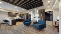 This well-appointed single-floor space blends contemporary design with functional living areas, creating an inviting and stylish environment throughout. The central lounge area serves as the heart of the space, anchored by a dramatic floor-to-ceiling stainless steel fireplace surround with a linear gas fireplace that adds both warmth and a bold visual statement. Exposed dark wood beam accents along the ceiling add architectural depth and contrast against the white upper surfaces, while recessed lighting ensures the space is well-lit throughout. Mid-century modern seating in rich teal blue with warm wood legs is arranged throughout the lounge, complemented by a sectional sofa positioned beside a large picture window that offers views of the surrounding neighborhood. The kitchen is fully equipped with dark espresso cabinetry, stainless steel appliances including a full-size refrigerator, range, and microwave, all set against crisp white quartz countertops. A peninsula island with bar seating provides additional workspace and casual dining. Light hardwood flooring in the kitchen transitions seamlessly to plush, textured carpet in the lounge and seating areas. Colorful artwork and thoughtful décor accents add personality, rounding out a space that is both functional and visually cohesive.