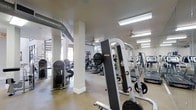 The Esplanade Fitness Center is a single-level facility designed to support a comprehensive workout experience. The open floor plan provides ample space for a wide variety of equipment, including treadmills, cable machines, weight stations, and cardio machines such as ellipticals and upright bikes. Strength-training apparatus, including a free-weight rack with plate-loaded machines, is positioned throughout the space for easy access and efficient use.

The interior features an industrial-inspired aesthetic, with exposed ductwork running along the ceiling and white structural columns that define zones within the gym without enclosing the space. Ceiling fans are installed overhead to promote air circulation, complementing the fluorescent lighting that keeps the facility well-lit throughout. Large windows along the perimeter invite natural light into the space, creating a bright and energizing atmosphere.

The flooring is finished in a durable, easy-to-maintain material suited for high-traffic fitness use. Wall-mounted mirrors enhance the sense of openness while allowing users to monitor their form. Overall, the Esplanade Fitness Center offers a functional and well-appointed environment for a wide range of fitness activities.