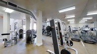 The Esplanade Fitness Center is a single-level facility designed to support a comprehensive workout experience. The open floor plan provides ample space for a wide variety of equipment, including treadmills, cable machines, weight stations, and cardio machines such as ellipticals and upright bikes. Strength-training apparatus, including a free-weight rack with plate-loaded machines, is positioned throughout the space for easy access and efficient use.

The interior features an industrial-inspired aesthetic, with exposed ductwork running along the ceiling and white structural columns that define zones within the gym without enclosing the space. Ceiling fans are installed overhead to promote air circulation, complementing the fluorescent lighting that keeps the facility well-lit throughout. Large windows along the perimeter invite natural light into the space, creating a bright and energizing atmosphere.

The flooring is finished in a durable, easy-to-maintain material suited for high-traffic fitness use. Wall-mounted mirrors enhance the sense of openness while allowing users to monitor their form. Overall, the Esplanade Fitness Center offers a functional and well-appointed environment for a wide range of fitness activities.