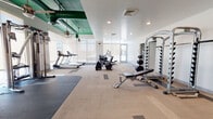 This fitness center is a thoughtfully designed workout facility that combines functionality with a modern industrial aesthetic. The space is defined by its striking green-painted exposed ductwork and structural elements along the ceiling, creating a bold visual contrast against the white walls and light herringbone-patterned flooring. Large floor-to-ceiling windows line one side of the facility, flooding the interior with abundant natural light and providing an open, airy atmosphere. Recessed lighting and ceiling fans complement the natural light throughout.

The facility is organized into distinct workout zones. The cardio area features a row of treadmills and elliptical machines positioned along the windowed wall, allowing users to exercise with a view. A dedicated strength training zone includes a multi-station cable machine, a power rack with a weight bench, and a comprehensive dumbbell rack. Additional equipment includes stationary and Stages cycling bikes, a medicine ball rack with an assortment of weighted balls, and an inversion table for stretching and recovery. Rubber flooring mats are strategically placed throughout to define workout zones and provide safety underfoot. A wall-mounted flat-screen TV is available for entertainment. Restroom facilities are accessible directly from the gym floor, adding convenience for users.