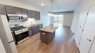 Located at 18400 Orchard Trail in Lakeville, Minnesota, this two-bedroom, one-bathroom unit offers a well-designed single-floor layout that makes efficient use of its space. The kitchen is a standout feature, showcasing rich dark cabinetry, striking granite countertops with bold veining, a subway tile backsplash, and a full suite of stainless steel appliances including a gas range, microwave, and dishwasher. A central kitchen island with matching granite provides additional prep and dining space, illuminated by modern pendant lighting overhead. Wood-style flooring flows throughout the main living and kitchen areas, lending warmth and continuity to the space. The primary bedroom is a comfortable, light-filled room with plush carpet, a ceiling fan, a window with outdoor views of surrounding greenery, and a closet. The second bedroom also features wood-style flooring and a window that brings in natural light. The bathroom includes a full tub and shower combination with clean white surrounds. A dedicated laundry area houses a stacked washer and dryer unit, adding everyday convenience. The overall aesthetic is clean and contemporary, with neutral tones and modern finishes throughout.