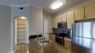 This single-floor residence offers a thoughtfully designed interior with notable attention to finish quality and layout. The kitchen serves as a central highlight, featuring a full suite of stainless steel appliances including a refrigerator, electric range with oven, and an over-the-range microwave. A double-basin sink is positioned on a kitchen island or peninsula with rich, dark granite-style countertops that add a sense of elegance to the space. The cabinetry is finished in a light wood tone, providing ample upper and lower storage throughout. A mosaic tile backsplash in earthy tones runs along the cooking wall, lending a contemporary and stylish detail to the kitchen. Crown molding accents the upper walls, adding a refined architectural touch. The ceilings are notably tall, contributing to an open and airy atmosphere. Adjacent to the kitchen, an arched passageway leads to a secondary area featuring built-in open shelving, ideal for storage or display. Wood-look flooring is visible in the neighboring space, complementing the overall warm, neutral color palette. Recessed lighting and flush-mount ceiling fixtures ensure the space is well-lit throughout.