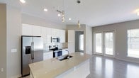 This single-floor 2-bedroom, 2-bathroom residence showcases a clean, modern aesthetic with thoughtfully designed living spaces. The open-concept kitchen is a standout feature, boasting high-gloss white cabinetry that reaches toward the ceiling, a suite of stainless steel appliances including a built-in microwave, electric range, and a full-size refrigerator. A generous kitchen island with a light quartz countertop and an undermount sink serves as both a functional workspace and a natural gathering point. Stylish pendant lights hang above the island, adding a refined touch to the space. The kitchen flows seamlessly into the living area, which is bathed in natural light from large glass French doors that open to the outdoors, creating an airy and connected atmosphere. Neutral-toned walls, recessed lighting, and smooth hardwood-style flooring unify the space with a cohesive, contemporary palette. The layout provides a practical and comfortable flow between rooms, making it well-suited for everyday living. With its modern finishes, abundant natural light, and open design, this residence offers a stylish and functional living environment.