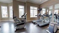 This single-level fitness space is designed to support an effective cardio-focused workout routine. The room is finished with warm taupe-toned walls accented by crisp white crown molding and window trim, lending a polished, upscale aesthetic. Large white-framed windows line the exterior wall, flooding the space with natural light and providing views of the surrounding outdoor greenery, creating an inviting atmosphere for exercise. The durable dark rubber flooring is both practical and visually grounding, offering stability underfoot during workouts. The equipment lineup includes multiple treadmills and an upright stationary bike, all from reputable commercial-grade brands, ensuring a quality exercise experience. A wall-mounted flat-screen television is positioned for easy viewing from the cardio machines, adding an entertainment element to workouts. A large mirror is affixed to one wall, visually expanding the space and allowing users to monitor their form. The overall ambiance is clean, bright, and well-maintained, reflecting a thoughtfully designed fitness amenity suited for regular use.