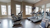 This single-level fitness space is designed to support an effective cardio-focused workout routine. The room is finished with warm taupe-toned walls accented by crisp white crown molding and window trim, lending a polished, upscale aesthetic. Large white-framed windows line the exterior wall, flooding the space with natural light and providing views of the surrounding outdoor greenery, creating an inviting atmosphere for exercise. The durable dark rubber flooring is both practical and visually grounding, offering stability underfoot during workouts. The equipment lineup includes multiple treadmills and an upright stationary bike, all from reputable commercial-grade brands, ensuring a quality exercise experience. A wall-mounted flat-screen television is positioned for easy viewing from the cardio machines, adding an entertainment element to workouts. A large mirror is affixed to one wall, visually expanding the space and allowing users to monitor their form. The overall ambiance is clean, bright, and well-maintained, reflecting a thoughtfully designed fitness amenity suited for regular use.