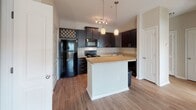 This single-floor residence in North Charleston, SC offers a well-designed, open-concept layout that seamlessly connects the main living areas. The kitchen is a standout feature, showcasing rich dark cabinetry with sleek hardware, granite-style countertops, a white subway tile backsplash, and a central island with pendant lighting overhead. A full suite of black appliances — including a refrigerator, electric range, microwave, and dishwasher — provides functional convenience. A decorative lattice cabinet adds a distinctive touch above the refrigerator. Warm hardwood-style flooring flows throughout the kitchen and main living areas, creating a cohesive and inviting aesthetic. The living space is open and airy, with crown molding accents and neutral walls that allow for versatile decorating. Natural light streams in through windows fitted with blinds, brightening the interior throughout the day. The bedroom is carpeted for comfort, features a ceiling fan, and receives generous natural light from a large window. The bathroom is cleanly appointed with a granite-topped vanity, dark cabinetry, and a full tub/shower combination. Multiple interior doors provide access to closets and additional storage, rounding out this practical and well-appointed North Charleston home.