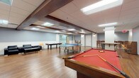 This single-floor recreational space is designed to serve as a multi-purpose amenity area, combining active game zones with relaxed lounge seating. The open layout accommodates a full-sized pool table with a bold red felt surface, a blue ping-pong table, and a foosball table, providing ample room for gameplay without crowding. Warm hardwood flooring runs throughout, contrasting pleasantly with the two-tone walls — a cool grey upper half above crisp white wainscoting — which give the space a clean, polished appearance.

Exposed dark wood beams add architectural character to the drop ceiling, which is fitted with recessed and fluorescent panel lighting to ensure even illumination across the entire area. A dedicated lounge nook is equipped with black leather sofas, a low coffee table, and wall-mounted flat-screen televisions, creating a comfortable viewing and socializing area. High-top bistro tables with wooden barstools are positioned along the perimeter, offering casual seating options.

Glass-paneled doors and white interior doors provide access to adjacent rooms, adding transparency and connectivity to the layout. The overall ambiance is casual yet well-appointed, making this space well-suited for social gatherings and recreational activities.