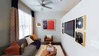 This studio presents a thoughtfully designed single-floor layout that maximizes every square foot of available space. The living area serves as the central hub, featuring rich hardwood-style flooring that anchors the room with warmth and durability. Neutral gray walls provide a versatile backdrop, allowing the curated artwork and décor accents to take center stage. A ceiling fan with a brushed metal finish adds both functionality and a contemporary touch overhead.

Natural light filters in through a large window dressed with layered blinds and patterned curtains, creating a bright yet controllable atmosphere. The living space comfortably accommodates a plush sofa, a leather accent chair, and a distinctive live-edge wood coffee table atop a shag area rug, highlighting the room's character. A wall-mounted flat-screen television is framed by a gallery-style arrangement of gold-framed artwork, adding an artistic focal point. An arched brass floor lamp introduces ambient lighting and a touch of elegance. The overall ambiance is modern and eclectic, blending artistic expression with practical, comfortable living in a compact, efficient footprint.