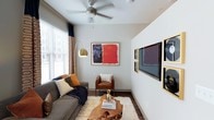 This studio presents a thoughtfully designed single-floor layout that maximizes every square foot of available space. The living area serves as the central hub, featuring rich hardwood-style flooring that anchors the room with warmth and durability. Neutral gray walls provide a versatile backdrop, allowing the curated artwork and décor accents to take center stage. A ceiling fan with a brushed metal finish adds both functionality and a contemporary touch overhead.

Natural light filters in through a large window dressed with layered blinds and patterned curtains, creating a bright yet controllable atmosphere. The living space comfortably accommodates a plush sofa, a leather accent chair, and a distinctive live-edge wood coffee table atop a shag area rug, highlighting the room's character. A wall-mounted flat-screen television is framed by a gallery-style arrangement of gold-framed artwork, adding an artistic focal point. An arched brass floor lamp introduces ambient lighting and a touch of elegance. The overall ambiance is modern and eclectic, blending artistic expression with practical, comfortable living in a compact, efficient footprint.