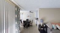 This single-floor residence offers a warm and comfortable living environment highlighted by an open-concept layout that seamlessly connects the living and dining areas. The dining space is anchored by a round dark-wood table paired with matching chairs, positioned beneath a ceiling fan with integrated lighting — a practical and stylish focal point of the room. Soft, neutral beige walls and light carpeted flooring create a calm, cohesive atmosphere throughout the space. Large windows dressed with decorative patterned curtains allow generous natural light to filter in, enhancing the airy feel of the interior. A textured popcorn ceiling adds a classic residential character overhead. A half-wall partition elegantly separates the dining area from the adjacent kitchen, which features white cabinetry and a bright, clean aesthetic. The living area flows naturally from the dining space, offering room for comfortable seating arrangements. Thoughtful decorative touches — including wall art and accent lamps — contribute to the welcoming ambiance of the home. The overall design prioritizes functionality and livability, making it a practical yet inviting single-level space.