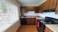 This single-floor residence at Heritage Green offers a well-organized layout with a practical mix of finishes throughout. The kitchen is a standout feature, designed in an L-shaped configuration with rich, dark wood cabinetry extending from floor to ceiling. Black appliances — including an electric range, dishwasher, and range hood — complement the neutral countertops, while a double-basin sink sits beneath a large window fitted with blinds, allowing natural light to brighten the space. Warm hardwood flooring runs through the kitchen and into the adjoining living and entry areas, providing a cohesive and easy-to-maintain surface. The living space is open and bright, with white walls, clean baseboards, and natural light filtering in through multiple windows. Decorative wall art adds subtle character to the otherwise neutral palette. The bedroom is carpeted for comfort and features a ceiling fan with a light fixture, a window overlooking the surrounding community, and ample natural light. The overall layout, as seen in the dollhouse view, is compact yet functional, with clearly defined zones for living, cooking, and sleeping, making efficient use of the available floor plan.