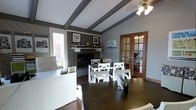 This thoughtfully designed single-floor space blends a professional and inviting atmosphere with classic architectural character. Exposed decorative ceiling beams add a distinctive structural charm, complementing the neutral gray and warm taupe color palette throughout. A ceiling fan provides both function and style overhead.

At the heart of the room sits a traditional fireplace with a detailed mantel, adorned with carefully selected décor and framed artwork, serving as a natural focal point. Dark hardwood floors ground the space with warmth and elegance, contrasting beautifully with the crisp white furniture.

The layout accommodates multiple workstations, including a white reception or service desk near the entrance and a central white dining or conference-style table with matching chairs, making the space highly functional for both collaborative and individual use. Natural light pours in through a large window, offering views of lush outdoor greenery.

A set of warm-toned wooden French doors with glass pane inserts connects this main area to an adjacent room, enhancing the sense of openness and flow. The overall ambiance is polished yet approachable, striking a balance between a professional work environment and a comfortable, homey setting.