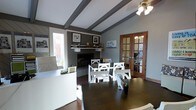 This thoughtfully designed single-floor space blends a professional and inviting atmosphere with classic architectural character. Exposed decorative ceiling beams add a distinctive structural charm, complementing the neutral gray and warm taupe color palette throughout. A ceiling fan provides both function and style overhead.

At the heart of the room sits a traditional fireplace with a detailed mantel, adorned with carefully selected décor and framed artwork, serving as a natural focal point. Dark hardwood floors ground the space with warmth and elegance, contrasting beautifully with the crisp white furniture.

The layout accommodates multiple workstations, including a white reception or service desk near the entrance and a central white dining or conference-style table with matching chairs, making the space highly functional for both collaborative and individual use. Natural light pours in through a large window, offering views of lush outdoor greenery.

A set of warm-toned wooden French doors with glass pane inserts connects this main area to an adjacent room, enhancing the sense of openness and flow. The overall ambiance is polished yet approachable, striking a balance between a professional work environment and a comfortable, homey setting.