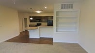 This single-floor space showcases an open-concept design that seamlessly connects the kitchen and living area. The kitchen is outfitted with dark espresso cabinetry, granite-style countertops, and black appliances, including a refrigerator, stove, and microwave. A peninsula breakfast bar extends from the kitchen, providing casual seating and serving as a natural divider between the kitchen and the adjacent living space. Pendant lighting above the peninsula adds a focused, functional accent to the area, complemented by recessed lighting throughout. A standout feature of the living area is the built-in wall shelving unit, offering generous display and storage space. The flooring transitions from dark hardwood in the kitchen and dining area to plush neutral carpet in the living space, creating a clear distinction between zones while maintaining a cohesive aesthetic. Ceiling vents are strategically placed for efficient climate control throughout the space. The overall ambiance is clean and functional, with a neutral color palette of white walls and dark cabinetry contrasts that lend the space a modern, well-organized feel suitable for everyday living.