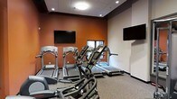 This fitness center is designed to provide a focused and functional workout environment. The space features a dedicated cardio area equipped with multiple treadmills and an elliptical machine, offering users a variety of options for aerobic exercise. Two wall-mounted flat-screen televisions are positioned strategically throughout the room, allowing users to stay entertained during their workouts. The room is finished with a durable, speckled fitness flooring that is both practical and easy to maintain. The walls feature a bold terracotta-orange accent paired with neutral tones, creating an energizing atmosphere that is conducive to physical activity. Recessed and flush-mount ceiling lighting provides bright, even illumination throughout the space. A window along one wall allows for a degree of natural light to enter, adding to the overall comfort of the environment. The drop-tile ceiling and clean architectural finishes give the space a polished, professional appearance. The layout is thoughtfully organized to maximize usable floor space while maintaining clear pathways between equipment, ensuring safe and comfortable use for all visitors.