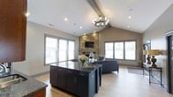 This single-floor space showcases a thoughtfully designed open-concept layout that seamlessly connects the kitchen and living areas into one cohesive, inviting environment. The vaulted ceiling, accented by a prominent exposed wood beam, lends a sense of grandeur and airiness throughout the main living space. A statement chandelier with candle-style lighting anchors the room and adds a warm, refined touch overhead.

The kitchen features a large central island topped with dark granite countertops, complemented by deep navy cabinetry with gold hardware — a sophisticated contrast that draws the eye. Recessed lighting throughout ensures the space is well-lit at all times.

The adjoining living area is anchored by a floor-to-ceiling natural stone fireplace, which serves as a dramatic architectural focal point. The stonework extends upward, adding texture and rustic character that balances the modern finishes found elsewhere in the space.

Large windows line the perimeter of the room on multiple sides, flooding the interior with abundant natural light and offering views of the surrounding exterior. Light hardwood flooring runs throughout, tying the kitchen and living zones together with warmth and continuity. The overall ambiance is a harmonious blend of modern elegance and rustic charm.