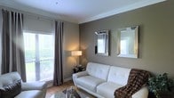 This single-floor space exudes a polished, contemporary aesthetic that balances comfort with refined style. The living area serves as the heart of the space, showcasing warm taupe-toned walls accented by crisp white crown molding that adds an elegant architectural detail. Large windows dressed with floor-length charcoal drapes and horizontal blinds allow an abundance of natural light to filter in, creating a bright and welcoming atmosphere with views of lush greenery outside.

The living room is thoughtfully appointed with a tufted white leather sofa, layered with a cozy textured throw, and complemented by a soft area rug and a sleek glass coffee table. A pair of mirrored, beveled-frame wall mirrors are strategically placed to reflect light and enhance the sense of depth and openness in the room. A stylish table lamp provides warm ambient lighting, adding to the inviting character of the space.

The hardwood flooring throughout lends warmth and continuity, tying the design elements together cohesively. Overall, the space presents a harmonious blend of modern furnishings and classic architectural touches, resulting in an interior that feels both sophisticated and livable.