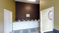 This communal laundry facility is designed with practicality and efficiency in mind. The single-floor space features a row of commercial-grade washers and dryers lined along a bold, dark brown accent wall, creating a clear visual anchor for the room. The machines are uniformly arranged for straightforward access, and an instructional notice board is mounted on the wall above, providing users with operating guidelines.

The room's color palette combines the deep brown feature wall with warm yellow-green tones on the adjacent walls, lending the space a welcoming feel despite its utilitarian purpose. The flooring consists of a durable, easy-to-clean tile in a neutral checkered pattern, well-suited to the demands of a high-traffic laundry environment.

Overhead fluorescent lighting ensures the space is well-lit throughout, supporting a functional and comfortable user experience. A doorway on the right side of the room provides access to an additional area, suggesting the facility may include supplementary amenities. The overall layout is open and uncluttered, making the space easy to navigate. Signage is clearly posted near the entrance, maintaining an organized and informative environment for all users.