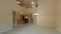 This two-story residence offers an open-concept design that maximizes the sense of space and flow throughout the main living areas. The living room is expansive, featuring impressively tall ceilings that extend upward to reveal a second-floor overlook, adding a dramatic architectural element to the interior. Soft, neutral-toned carpeting covers the main floor, contributing to a warm and inviting ambiance. A chandelier with multiple light fixtures serves as a focal point, providing both style and illumination to the open space. The kitchen is accessible directly from the living area and is outfitted with warm wood-toned cabinetry, stainless steel appliances including a refrigerator and gas range, and dark countertops. A kitchen island provides additional workspace and storage. An interior staircase connects the main floor to the upper level, where a landing overlooks the living space below. The upper floor features its own recessed lighting and railing, reinforcing the home's open vertical design. Neutral wall tones throughout complement the overall layout, creating a cohesive and functional living environment across both floors.